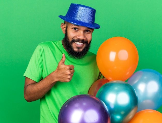 Jovem afro-americano sorridente com chapéu de festa segurando balões aparecendo o polegar isolado na parede verde