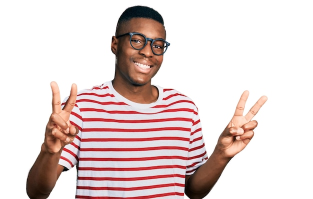 Jovem afro-americano vestindo camiseta casual e óculos sorrindo olhando para a câmera mostrando os dedos fazendo o sinal de vitória número dois