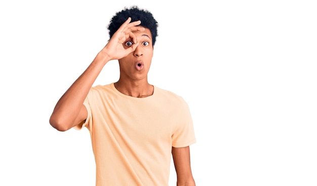 Foto jovem afro-americano vestindo roupas casuais fazendo ok gesto chocado com o rosto surpreso olho olhando por entre os dedos expressão incrédula