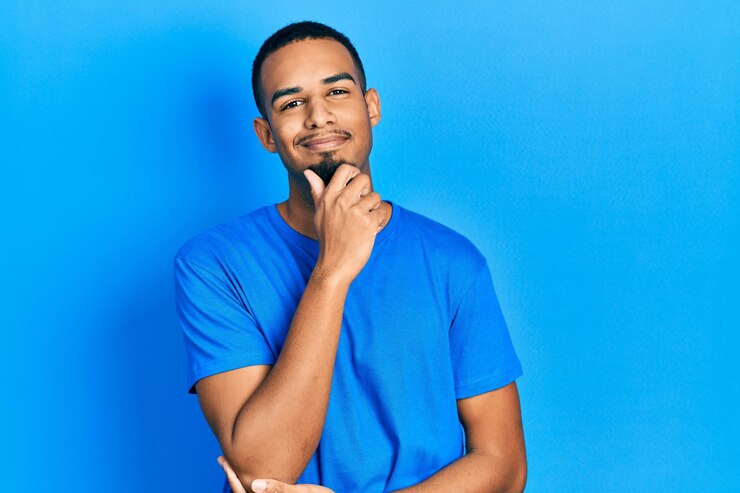 Foto jovem afro-americano vestindo uma camiseta azul casual olhando com confiança para a câmera com um sorriso com os braços cruzados e a mão levantada no queixo pensando positivo
