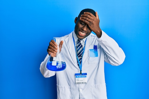 Jovem afro-americano vestindo uniforme de cientista segurando tubo de ensaio estressado e frustrado com a mão na cabeça, rosto surpreso e zangado