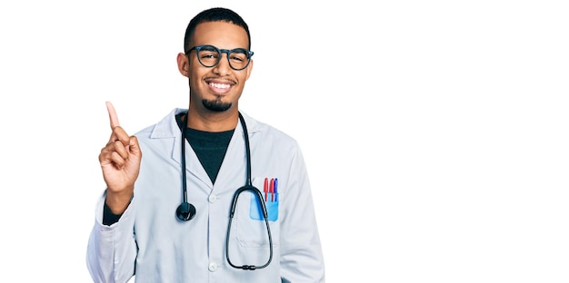 Jovem afro-americano vestindo uniforme de médico e estetoscópio apontando o dedo para cima com uma ideia de sucesso saindo e feliz número um