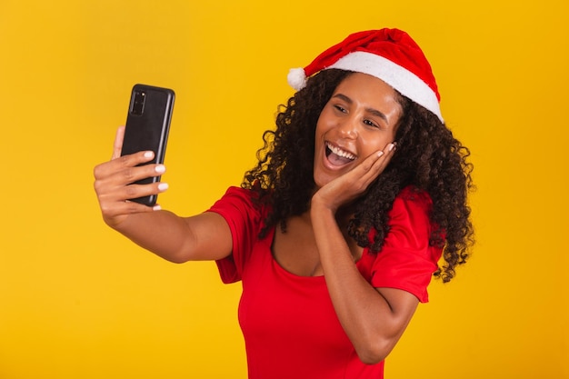 Jovem afro com chapéu de natal, fazendo videochamada em fundo amarelo.