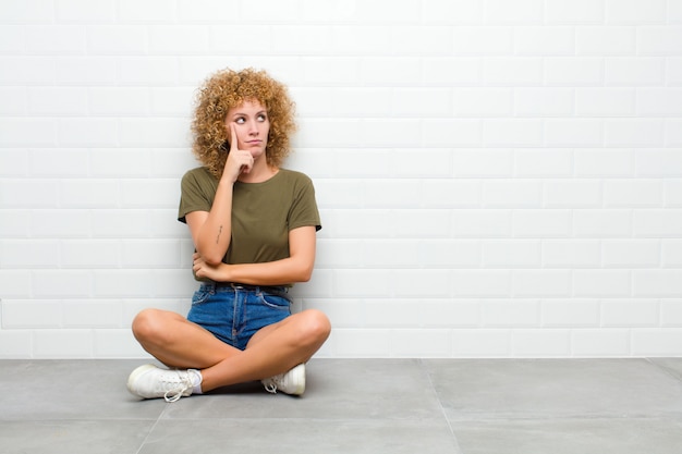 Jovem afro com um olhar concentrado, pensando com uma expressão duvidosa, olhando para cima e para o lado, sentada no chão