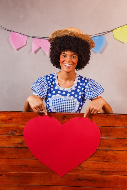 Jovem afro vestida para festa junina atrás de uma placa de madeira com um coração