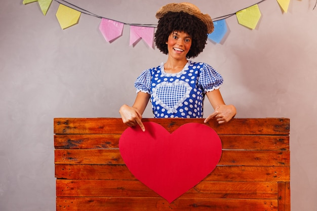 Jovem afro vestida para festa junina atrás de uma placa de madeira com um coração