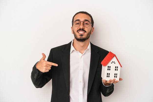 Jovem agente imobiliário segurando uma casa modelo isolada no fundo branco pessoa apontando com a mão para um espaço de cópia de camisa, orgulhoso e confiante