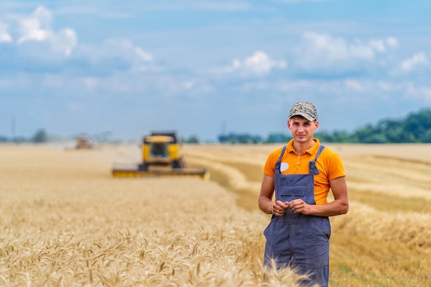 Jovem agricultor atraente em pé no campo de trigo. colheitadeira trabalhando no campo de trigo no fundo.