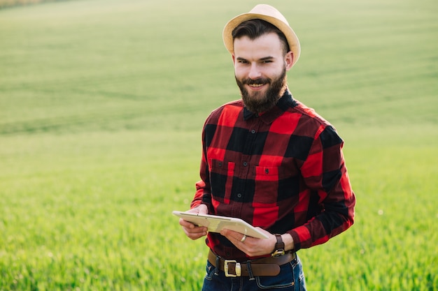 Jovem agricultor barbudo bonito com pé de tablet no campo de trigo no início do verão