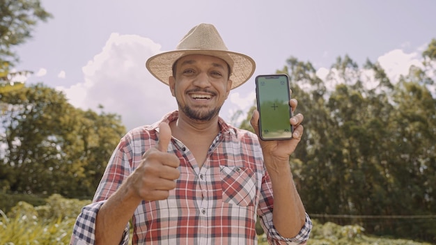 Jovem agricultor de chapéu sorrindo para a câmera e mostrando smartphone com tela de croma chave