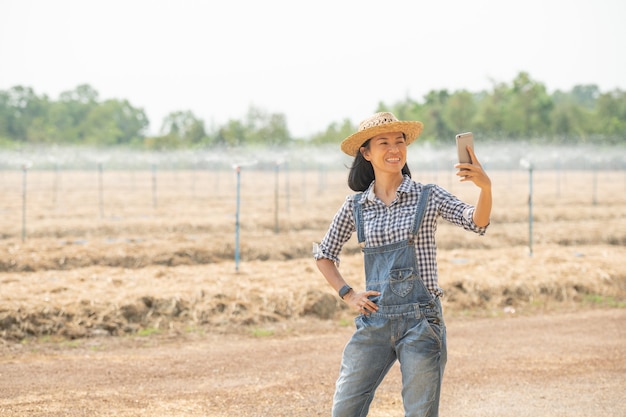 Jovem agricultor feminino da ásia em pé de chapéu na mulher do campo usando a tecnologia do telefone móvel para inspecionar no jardim agrícola. crescimento da planta. ecologia de conceito, transporte, ar puro, comida, produto biológico