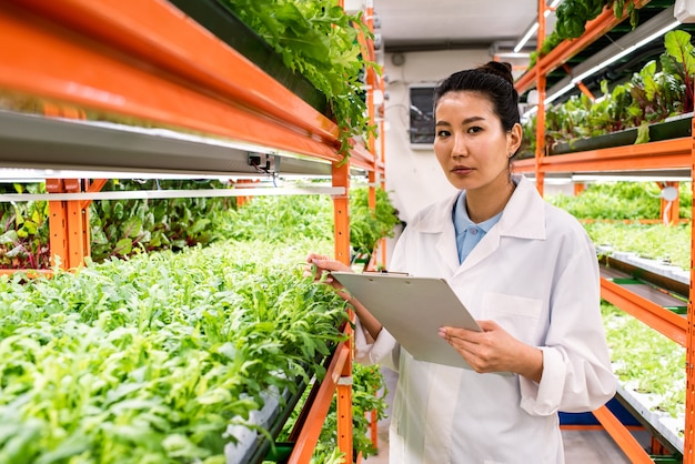 Jovem agrônomo contemporâneo de etnia asiática segurando documento enquanto faz pesquisas em estufa