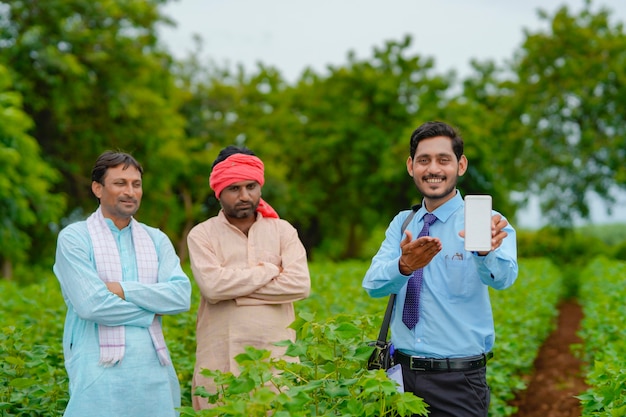 Jovem agrônomo indiano ou banqueiro apresentando smartphone com agricultores no campo de agricultura.