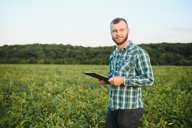 Jovem agrônomo segura tablet touch pad computador no campo de soja e examina as culturas antes da colheita Engenheiro agrônomo do conceito de agronegócios em um campo de soja com um tablet no verão