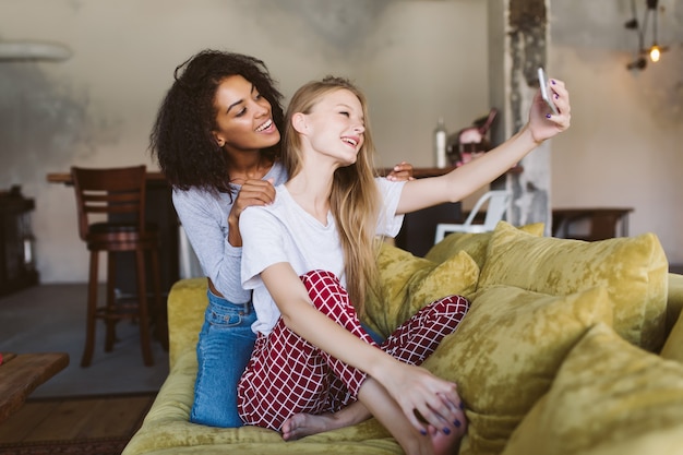 Foto jovem alegre afro-americana com cabelo escuro e encaracolado e uma linda mulher com cabelo loiro tirando fotos com alegria no celular em uma casa aconchegante