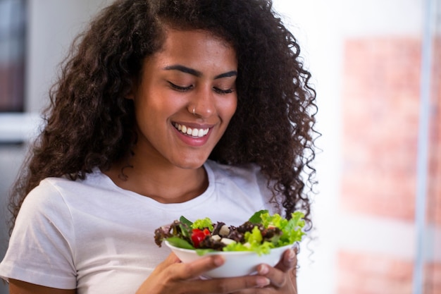 Foto jovem alegre afro-americana comendo salada de legumes na cozinha de casa