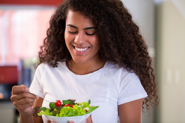 jovem alegre afro-americana comendo salada de legumes na cozinha de casa