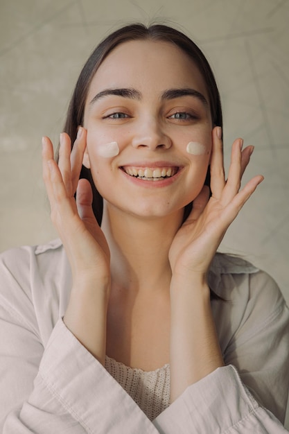 jovem alegre aplicando creme facial e olhando para a câmera enquanto faz a rotina de cuidados com a pele