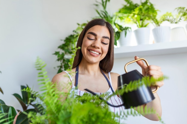 Jovem alegre aproveita o tempo em casa e rega a planta perto da janela em casa. mulher cuida de sua água de samambaia na árvore em um dia relaxante no jardim em casa.