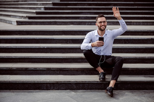 Jovem alegre barba por fazer elegantemente vestido, sentado na escada ao ar livre com fones de ouvido nas orelhas, segurando o telefone inteligente e acenando para um amigo.