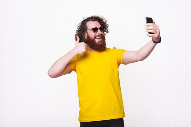 Jovem alegre barbudo está fazendo uma selfie em um estúdio.