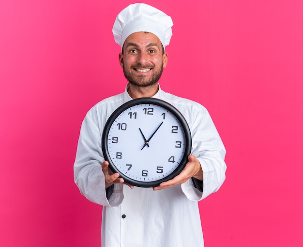 Jovem alegre, caucasiano, cozinheiro, com uniforme de chef e boné, olhando para a câmera, esticando o relógio em direção à câmera, isolada na parede rosa