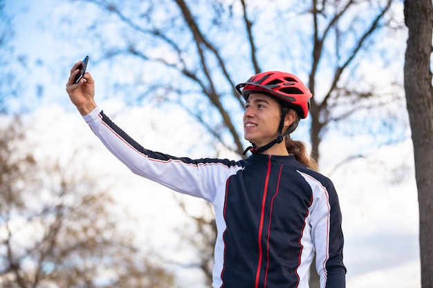 Jovem alegre ciclista masculina no sportswear e capacete tomando selfie em smartphone no parque