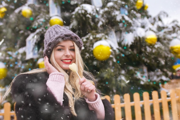 Jovem alegre com balas de chocolate caminhando perto da árvore do ano novo durante a queda de neve