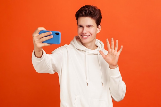 Jovem alegre com capuz elegante dizendo oi fazendo videochamada usando smartphone sorrindo com sorriso dentuço Conexão 4g Estúdio interno isolado em fundo laranja