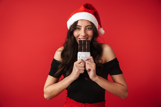 Jovem alegre com chapéu de Natal em pé, isolada sobre o vermelho, comendo chocolate