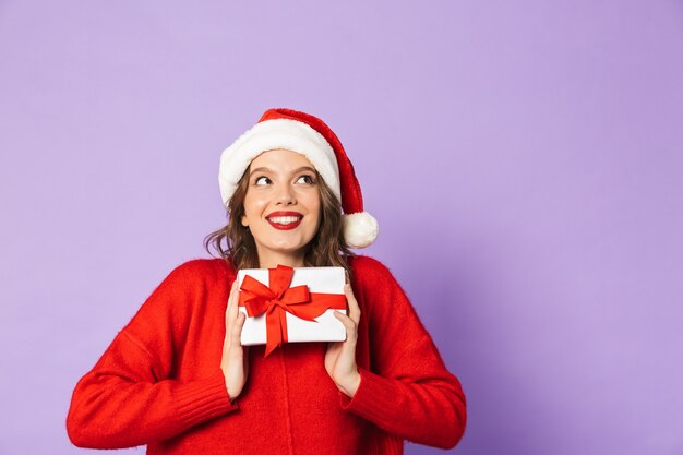 Jovem alegre com chapéu de Natal vermelho, isolada na parede violeta, segurando uma caixa de presente