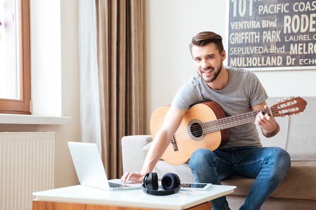 Jovem alegre com guitarra escrevendo música usando o laptop em casa