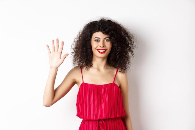 Jovem alegre com maquiagem e vestido vermelho, mostrando cinco dedos e sorrindo, em pé sobre um fundo branco. copie o espaço