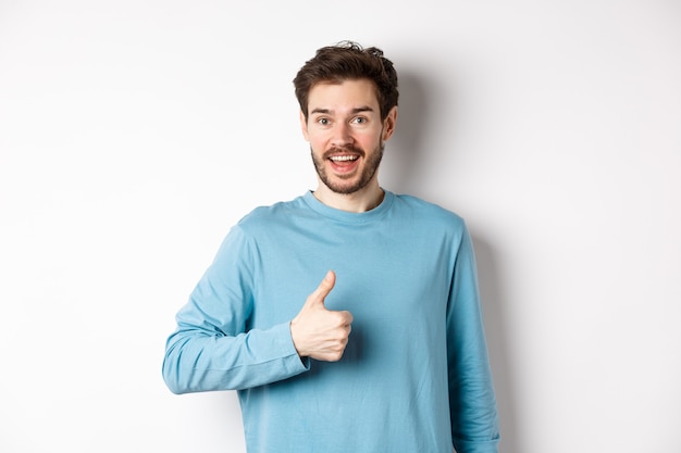 Jovem alegre concorda e diz que sim, mostra o polegar em aprovação, sorrindo feliz para a câmera, em pé sobre um fundo branco