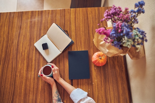 Jovem alegre, confiante e feliz, segurando uma xícara de chá em uma mesa com um caderno e flores