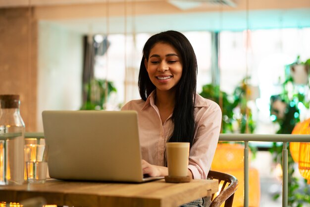 Jovem alegre de cabelos escuros sentada no café sorrindo enquanto usa o laptop e toma um copo de café com leite ao seu lado