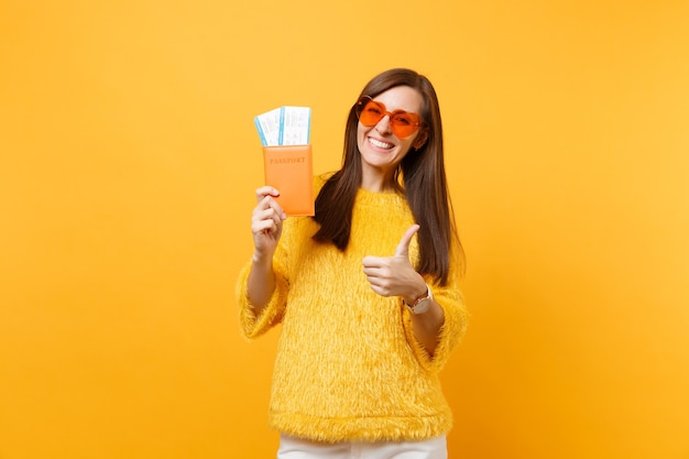 Jovem alegre de óculos coração laranja aparecendo o polegar, segurando o passaporte e os bilhetes de embarque isolados em fundo amarelo brilhante. Pessoas sinceras emoções, estilo de vida. Área de publicidade.