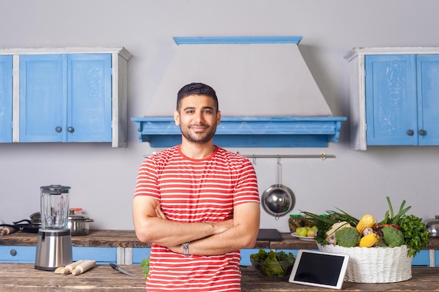 Jovem alegre de pé com as mãos cruzadas na cozinha moderna, olhando para a câmera com o tablet pc de sorriso e legumes verdes frescos na mesa atrás de receitas de nutrição vegetariana na internet