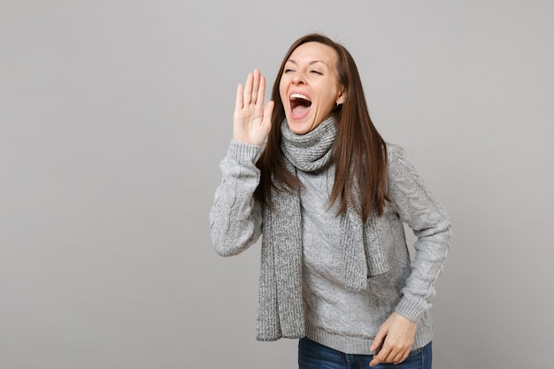 Jovem alegre de suéter cinza, lenço gritando com gesto de mão isolado no fundo da parede cinza. Estilo de vida de moda saudável, emoções sinceras de pessoas, conceito de estação fria. Simule o espaço da cópia.