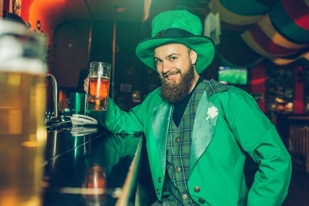 Foto jovem alegre e agradável no terno do st. patrick sente-se no balcão de bar no pub e segure a caneca de cerveja. ele olha e sorri. cara feliz.