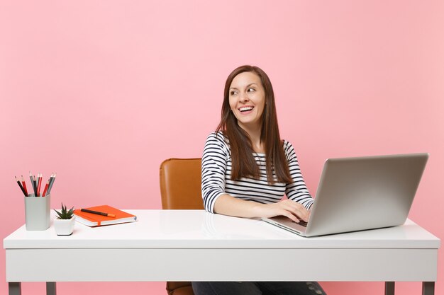 Jovem alegre e feliz olhando para o lado, trabalhando no projeto, enquanto está sentado no escritório com o laptop do pc