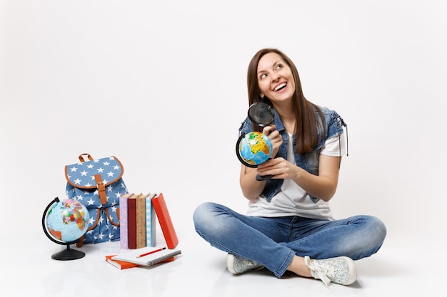 Jovem alegre e linda estudante segurando o globo do mundo e a lupa, sentada perto da mochila, livros escolares isolados na parede branca