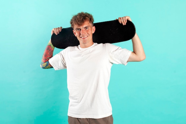 Jovem alegre e tatuado de camisa branca segurando um skate e sorrindo em fundo azul