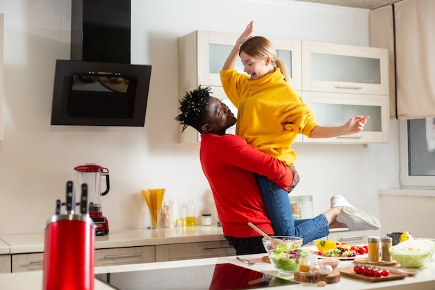 Jovem alegre em pé na cozinha sorrindo enquanto colocava sua namorada feliz no ar