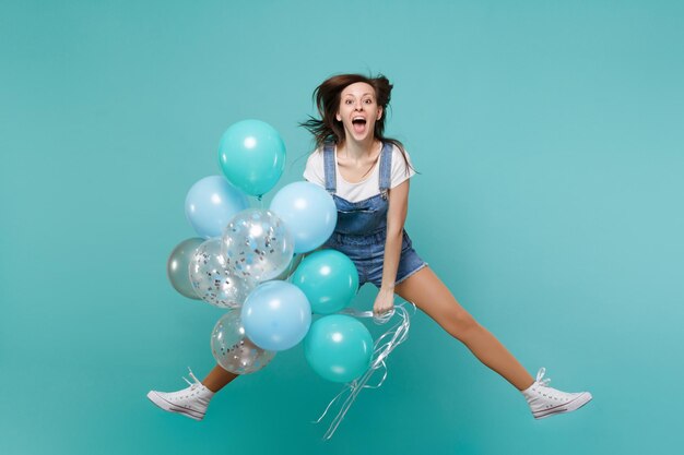 Jovem alegre em roupas jeans, mantendo a boca aberta, pulando alto, comemorando, segure balões de ar coloridos isolados em fundo azul turquesa. festa de aniversário, conceito de emoções de pessoas.