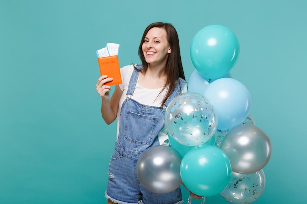 Jovem alegre em roupas jeans, segurando o bilhete de embarque do passaporte comemorando com balões de ar coloridos isolados no fundo azul turquesa. festa de aniversário, conceito de emoções de pessoas.