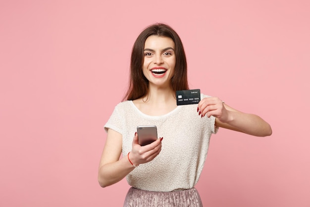 Jovem alegre em roupas leves casuais posando isolado no fundo rosa pastel. Conceito de estilo de vida de pessoas. Mock up espaço de cópia. Usando o telefone celular, digitando mensagem sms, segurando o cartão bancário de crédito.