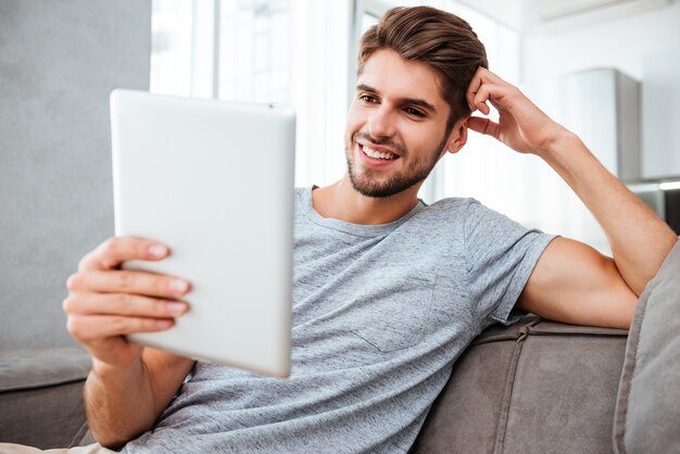 Jovem alegre em t-shirt cinza, sentado no sofá em casa enquanto olha no tablet.