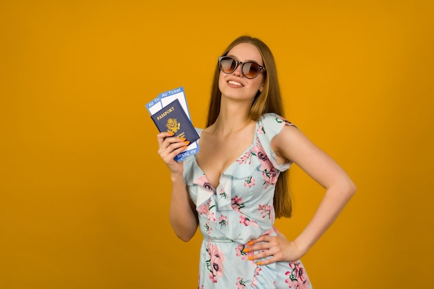 Jovem alegre em um vestido azul com flores e óculos de sol está segurando as passagens aéreas com um passaporte com um fundo amarelo. Alegra-se com a retomada do turismo após a pandemia do coronovírus.