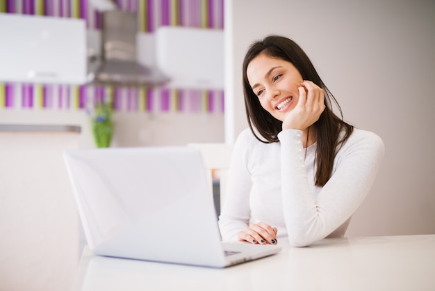 Jovem alegre está apoiando o cotovelo em uma mesa e segurando o queixo enquanto estiver usando um laptop.